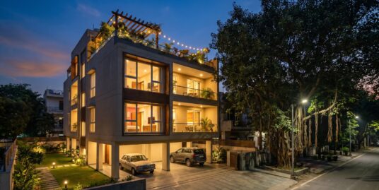 Best floor in a builder floor — a stilt-plus-four South Delhi building at blue hour with a ground-floor lawn and a lit terrace garden on the roof