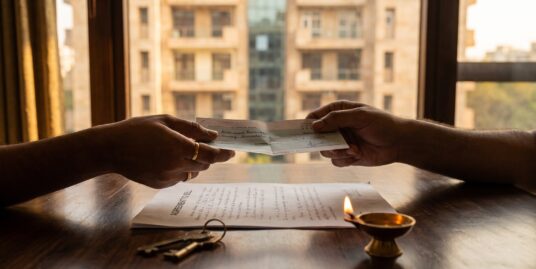 Token money in a property deal — a cheque handed across an agreement to sell with house keys beside it, a South Delhi builder floor in the evening light outside
