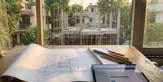 Building plan sanction in Delhi — an architect's blueprint of a stilt plus four floor building with a South Delhi rebuild under construction outside