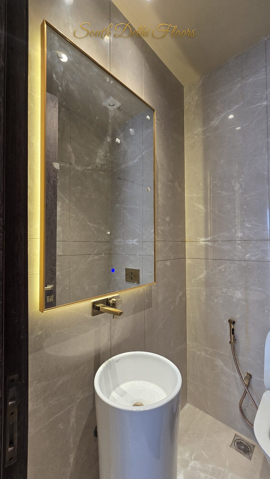 Powder room with freestanding pedestal basin, gold wall-mounted tap and backlit mirror on grey stone at an Indus Homes builder floor in GK-1, South Delhi