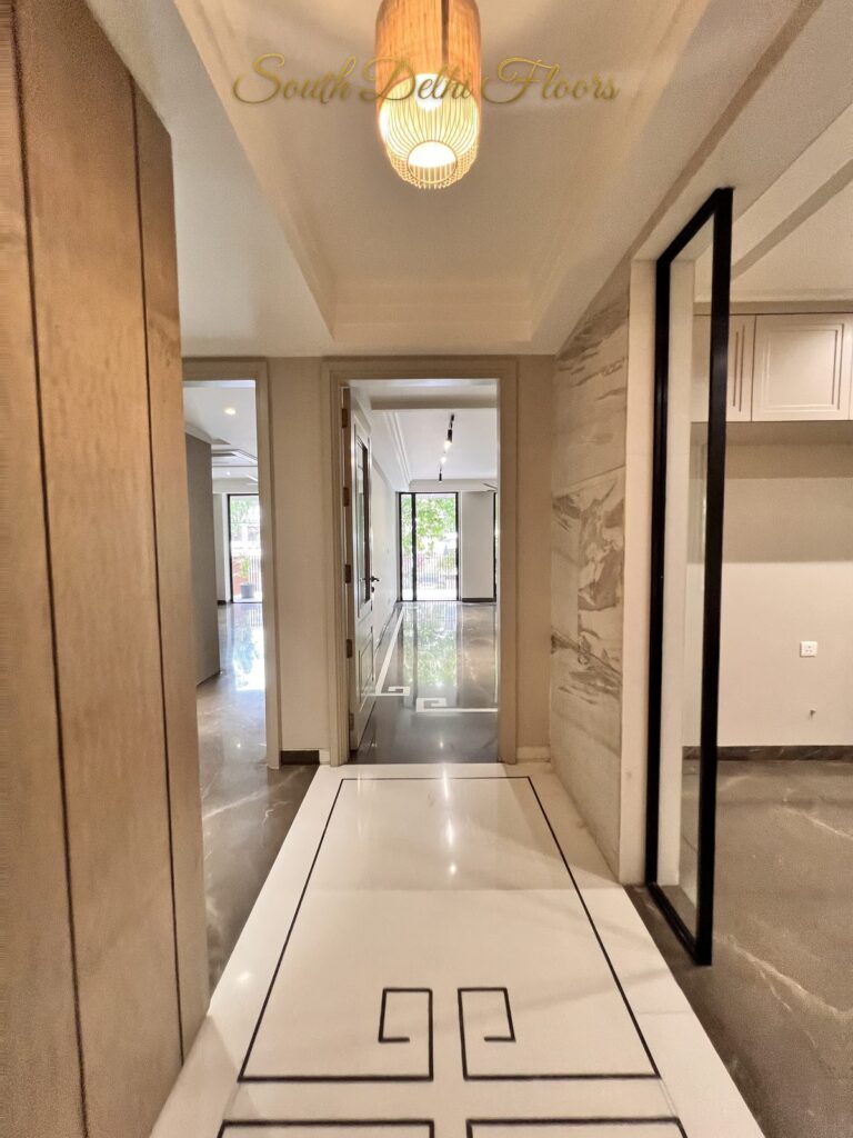 Black-framed glass pivot door opening off a marble corridor with a Greek key inlay at a Katarias luxury home in South Delhi by builder Sandeep Kataria