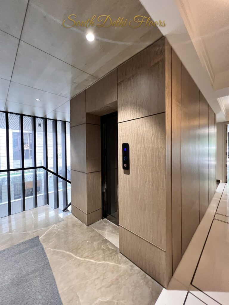 Elevator foyer with a veneer lift portal and doormat beside a black-framed glass stairwell at a Katarias luxury builder floor in South Delhi by Sandeep Kataria
