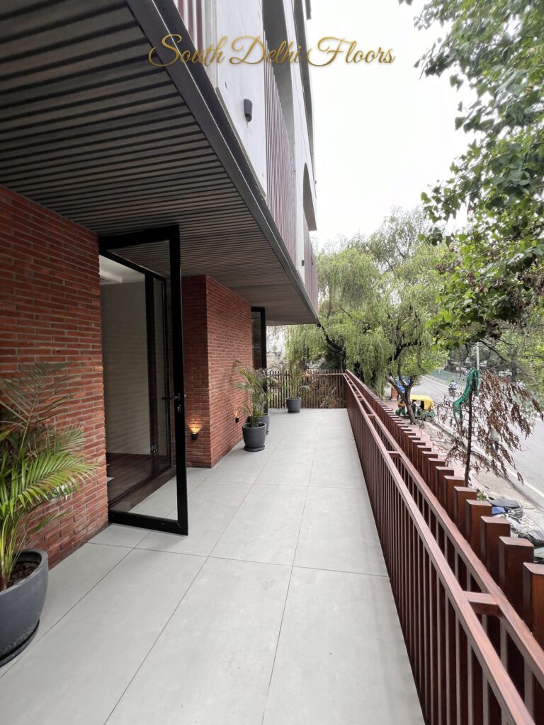 Front balcony with an exposed brick wall, black-framed glass door and terracotta slatted railing at a Katarias luxury home in South Delhi by builder Sandeep Kataria