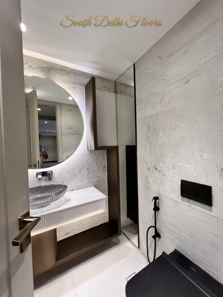 Powder room with a backlit round mirror on a textured stone wall above a fluted vanity at a Katarias luxury home in South Delhi by builder Sandeep Kataria