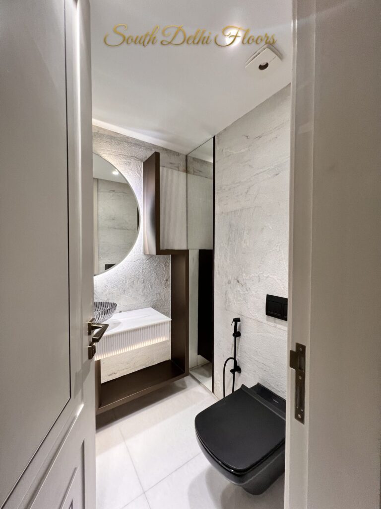 Powder room with a black wall-hung WC, white stone walls and a round backlit mirror at a Katarias luxury residence in South Delhi by developer Sandeep Kataria