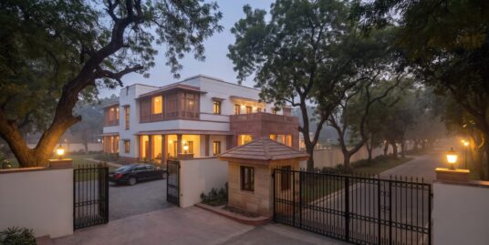 Renting property to embassy in Delhi — a gated residence on a tree-lined avenue with a sentry cabin at dusk