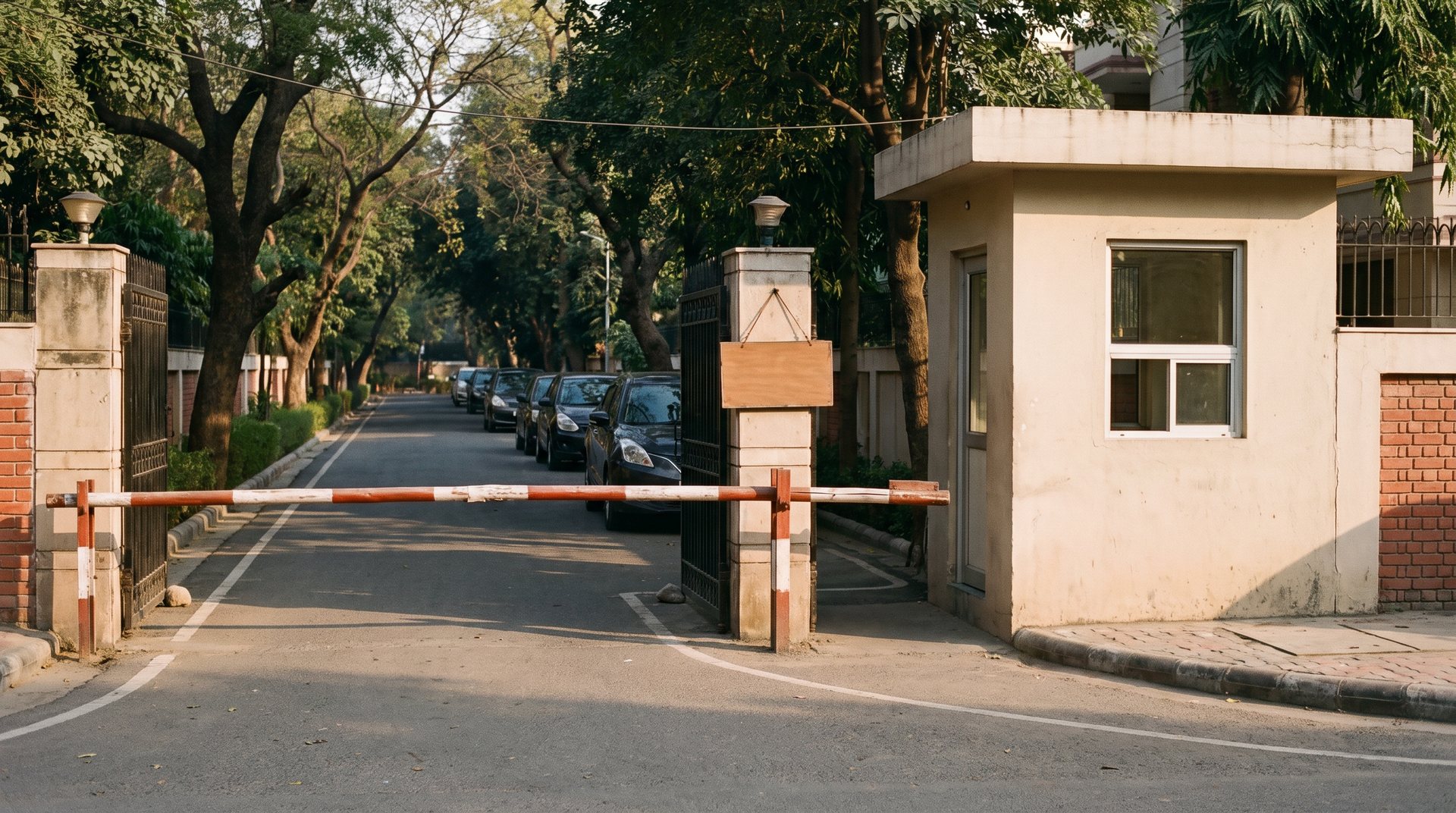 The gate of an upscale South Delhi residential colony with a boom barrier and guard cabin in afternoon light