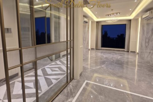 Living room seen through a bronze-framed glass partition in the Vasant Vihar D Block homes, with marble floors and a chandelier