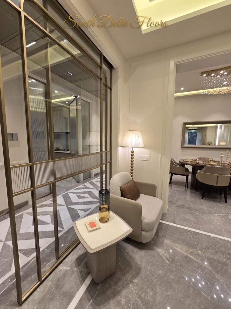 Furnished sitting corner behind a brass-framed glass partition at a Windchimes Construction floor in South Delhi, photographed by day